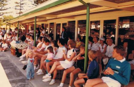 Members seated along the shaded clubhouse verandah watching tennis.
